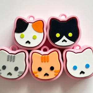 May include: Five cat-shaped charms with pink borders. Each charm features a different cat face design, including calico, black and white, gray, orange tabby, and white. The charms have a small loop at the top for attaching to keychains or other accessories.