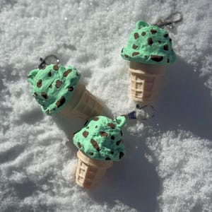 May include: Three mint chocolate chip ice cream cone keychains. The ice cream is light green with brown chocolate chips. The cones are beige with a waffle pattern. Each keychain has a silver clasp.