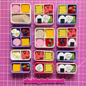 May include: Overhead shot of nine miniature bento boxes, each with different food items. The boxes are white, pink, and purple, and contain items like crackers, eggs, and rice balls. The background is a pink grid.