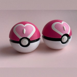 Puede incluir: Dos Poké Balls rosas y blancas con un diseño de corazón. La mitad superior es rosa con un corazón blanco, y la mitad inferior es blanca con una banda negra. Un botón blanco está en el centro.
