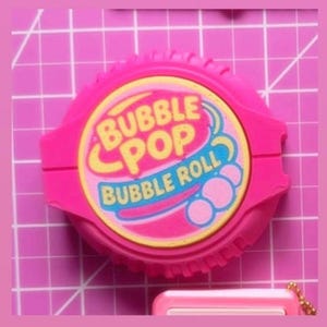 May include: A bright pink Bubble Pop Bubble Roll container. The container is round with a ridged edge and a rectangular section on each side. The label features the words "Bubble Pop" in yellow and "Bubble Roll" in blue.