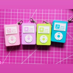 May include: Four colorful keychain accessories in pink, purple, green, and blue, each resembling a small music player. The pink one says "LOVE", the purple one says "VIBES", the green one says "LUCK", and the blue one says "CHILL".
