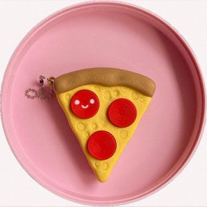 Może przedstawiać: Żółty brelok do kluczy w kształcie kawałka pizzy z brązową skórką i trzema czerwonymi pepperoni. Kawałek pizzy ma uśmiechniętą buzię i leży na różowym talerzu. Brelok jest przymocowany do srebrnego łańcuszka.