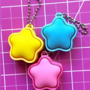 May include: Three star-shaped keychains in yellow, light blue, and pink, each featuring a silver-toned metal ball chain. The keychains are positioned on a cutting mat with visible white grid lines and numbers, showing a scale of approximately 3 inches.