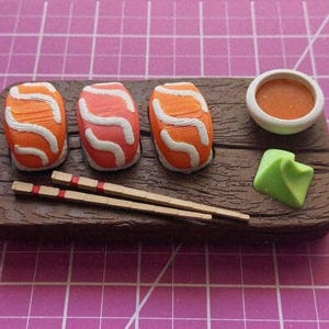 May include: Miniature sushi set on a wooden tray. The set includes three pieces of sushi with orange and white details, a small bowl of orange sauce, and a green wasabi piece. Two chopsticks are also included.