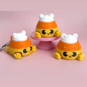 Puede incluir: Tres figuras con forma de caramelo de maíz con rasgos de oso. Las figuras son amarillas, naranjas y blancas, con ojos negros y una boca sonriente. Una figura está unida a un llavero. Una figura está sobre un pedestal rosa.