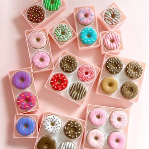 May include: A selection of colourful donuts in pink boxes is displayed from above. The donuts have various toppings and colours, including pink, blue, green, brown, and white. The boxes are arranged on a light pink surface, creating a visually appealing display.