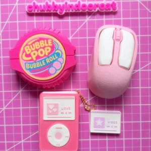 May include: A collection of pink novelty items, including a bubble gum roll, a computer mouse, a music player, and a small card with the word "Vibes". The bubble gum roll has the text "Bubble Pop Bubble Roll". The items are arranged on a pink grid.