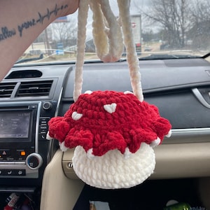 May include: A crocheted mushroom-shaped purse with a white strap. The purse is red with white accents and has a white crocheted base.