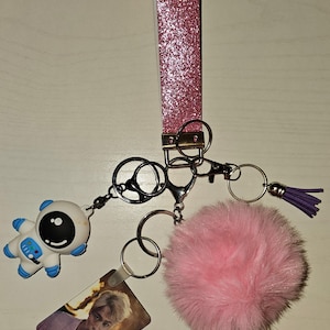 May include: Pink glitter keychain with a pink fluffy pom pom, a silver astronaut charm, and a photo card with a person on it.