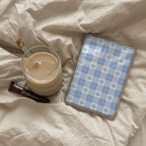 May include: A glass of coffee with a straw, a blue and white patterned tablet case, and a tube of rhode lip treatment on a beige bedspread. The straw has a small teddy bear decoration.