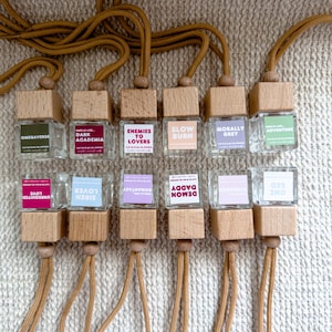 May include: A collection of small, clear glass bottles with wooden tops and tan cords. Each bottle has a label with text such as "OMEGAVERSE", "ENEMIES TO LOVERS", and "ONE BED". The bottles are arranged in rows.