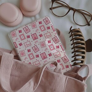 May include: A pink phone case with a repeating pattern of pink postage stamps, locks, and bows. The case is inside a pink tote bag. Other items include pink headphones, brown eyeglasses, and a brown hair clip.