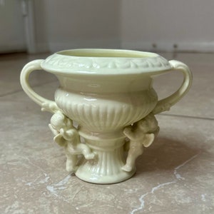May include: A small, white ceramic urn with two cherub handles. The urn has a fluted design and a base with a decorative scroll.