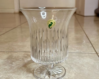May include: A clear glass vase with a cut glass design and a gold label with a green seahorse logo that reads "Waterford Crystal".