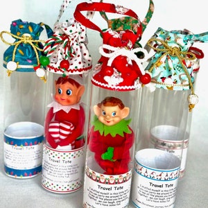 May include: Four clear plastic tubes with red and white fabric bags on top. Each tube contains a small elf doll. The tubes have labels that say "Travel Tote" and include a short poem about the elf.