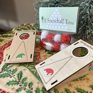 Peut inclure: Un jeu de lancer de boules de neige Elf en bois. L'ensemble comprend deux rampes en bois avec un trou circulaire, un sac de pompons rouges et blancs et une enseigne avec l'inscription "Elf Snowball Toss". Le jeu est posé sur une nappe festive sur le thème des fêtes.