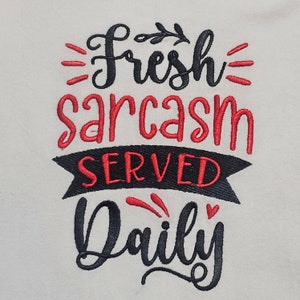 May include: Embroidered design on a white fabric background. The design says "Fresh Sarcasm Served Daily" in black and red lettering.