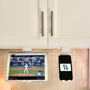 May include: White tablet and smartphone holders mounted under a kitchen cabinet. A tablet displays a baseball game, and a smartphone shows the text "CL 3Q". A small lamp and a potted plant are also visible.