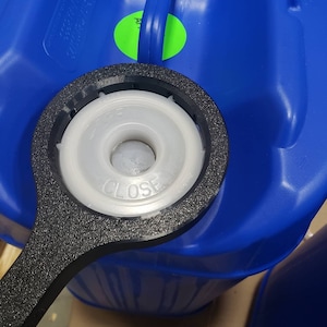 May include: A black plastic bung wrench positioned over the white circular cap of a large blue plastic container. The cap features moulded text that reads CLOSE and OPEN with directional arrows. A bright green circular sticker is visible on the container.