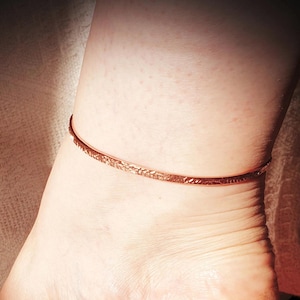 May include: A copper-coloured, textured anklet. The thin, hammered metal band encircles the ankle. The anklet has a simple, elegant design, reflecting light. The jewellery is a delicate accessory.
