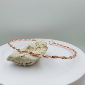 May include: A rose gold-coloured metal bracelet featuring a twisted wire design. The bracelet is an open-cuff style with a small circular loop at one end. The metal has a polished, reflective finish that catches the light along its spiraled surface.