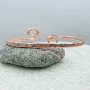 Peut inclure: Un bracelet manchette de couleur cuivre avec une texture martelée et un motif en spirale à une extrémité. Le bracelet est présenté sur une pierre grise, mettant en valeur ses détails artisanaux. Ce bijou est un accessoire élégant.