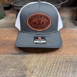 May include: Gray and white trucker hat with a brown leather patch featuring a stylized longhorn steer and floral design. The patch has white stitching around the edge. The hat has a Richardson 112 Original Trucker label on the back.