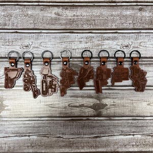 May include: Ten leather keychains with different construction vehicle designs. Each keychain is brown and features a silver metal ring.