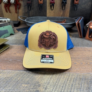 May include: A yellow and blue trucker hat with a brown leather patch that says "Cowgirl Up". The hat is adjustable and has a Richardson brand label.