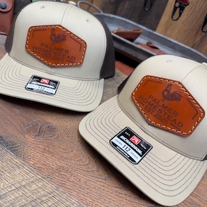 May include: Two brown and tan trucker hats with a brown leather patch on the front. The patch has a rooster design and the text "Palmer Homestead Est. 2025".