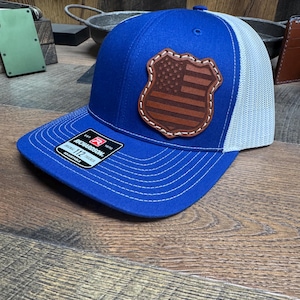 May include: A blue and white trucker hat with a brown leather patch featuring an American flag. The patch is stitched around the edges. The hat is adjustable and has a "Richardson" label.