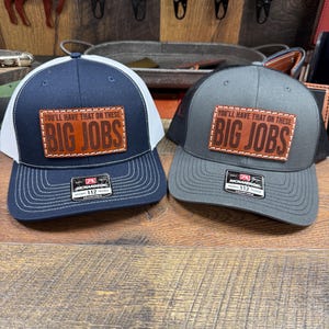 May include: Two trucker hats with a brown leather patch that says "You'll have that on these BIG JOBS". The hats are one navy blue and one gray.