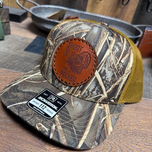 May include: A camouflage trucker hat with a brown leather patch featuring a turkey graphic and the text "SHOOT THE PECKER". The hat has a brown mesh back and a curved brim. The tag reads "RICHARDSON 112 TRUCKER".