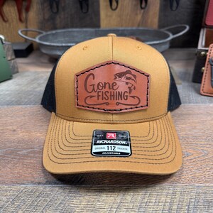 May include: A brown and black trucker hat with a brown leather patch that says "Gone Fishing" with a fish graphic. The hat is adjustable and has a black mesh back.