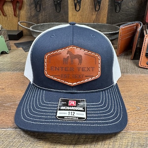 May include: A navy blue and white trucker hat with a brown leather patch featuring a horse and foal silhouette. The patch has the text "ENTER TEXT - EST. TEXT" on it. The hat is adjustable and has a Richardson 112 label.