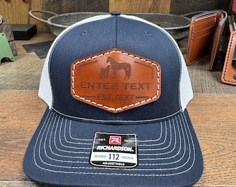 May include: A navy blue and white trucker hat with a brown leather patch featuring a horse and foal silhouette. The patch has the text "ENTER TEXT - EST. TEXT" on it. The hat is adjustable and has a Richardson 112 label.