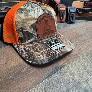 May include: A camouflage and orange mesh trucker hat with a brown leather patch that says "Legendary Turkey Hunter". The hat is made by Richardson and is adjustable.