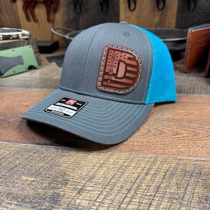 May include: Gray trucker hat with a blue mesh back and brim. The hat features a leather patch with the word "DURAMAX" and a design. The hat has blue stitching and a "RICHARDSON 112" tag.