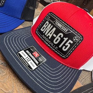 May include: A red, white, and black trucker hat with a black patch that says "The Volunteer State Tennessee BNA-615 Nashville". The hat is made by Richardson and is adjustable.