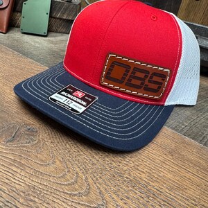 May include: A red, white, and navy blue trucker hat with a leather patch that reads "OBS". The hat has a mesh back and a curved brim. The brand is Richardson 112.