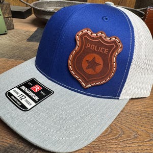 May include: A blue and gray baseball cap with a brown leather police badge patch. The patch has a star and the word "POLICE" stitched on it. The cap is adjustable and has a mesh back.