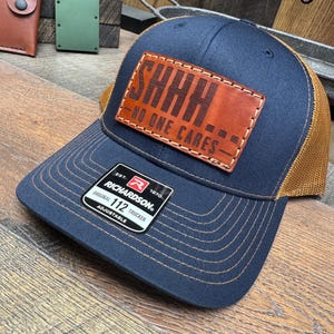 May include: A navy blue and brown trucker hat with a brown leather patch that says "SHHH - NO ONE CARES -" in brown lettering. The hat is adjustable and has a Richardson 112 label.