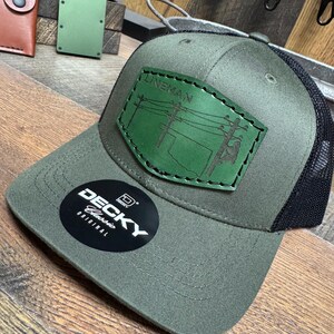 May include: Green and black baseball cap with a leather patch that says "Lineman" and depicts a lineman working on a power line. The cap is a Deckys Classic Original.
