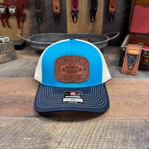 May include: A blue and white trucker hat with a navy blue brim. The hat features a brown leather patch with the words "Buckle Bunny" and decorative elements. The hat is adjustable and has a Richardson tag.