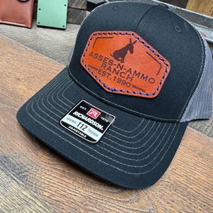 May include: Black trucker hat with a gray mesh back and a curved brim. The hat features a brown leather patch with the text "ASSES-N-AMMO RANCH EST. 1990" and a donkey silhouette. The hat is adjustable and made by Richardson.