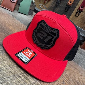 May include: A red flat bill snapback hat with a black mesh back. The hat features a black patch with the text "EMRY 21". A white and black tag reads "RICHARDSON FLAT BILL SNAPBACK EST. 1970".