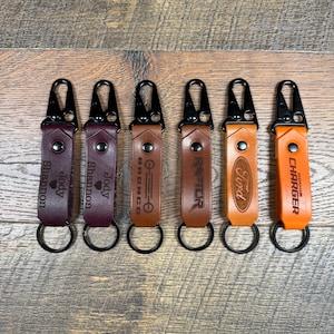 May include: Six leather keychains with different designs and names. The keychains are brown, burgundy, and orange. The names on the keychains are "Jody", "Shannon", "Bronco", "Raptor", "Ford", and "Dodge Charger".