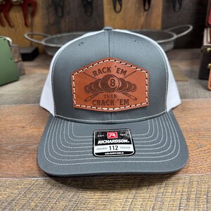 May include: Gray and white trucker hat with a brown leather patch that says "Rack 'Em Then Crack 'Em" with a pool ball and crossed pool sticks. The hat is adjustable and has the Richardson logo.