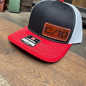 May include: A black, red, and white trucker hat with a red brim and a black crown. The hat features a brown leather patch with the text "C/10". The hat is adjustable and has a white mesh back.